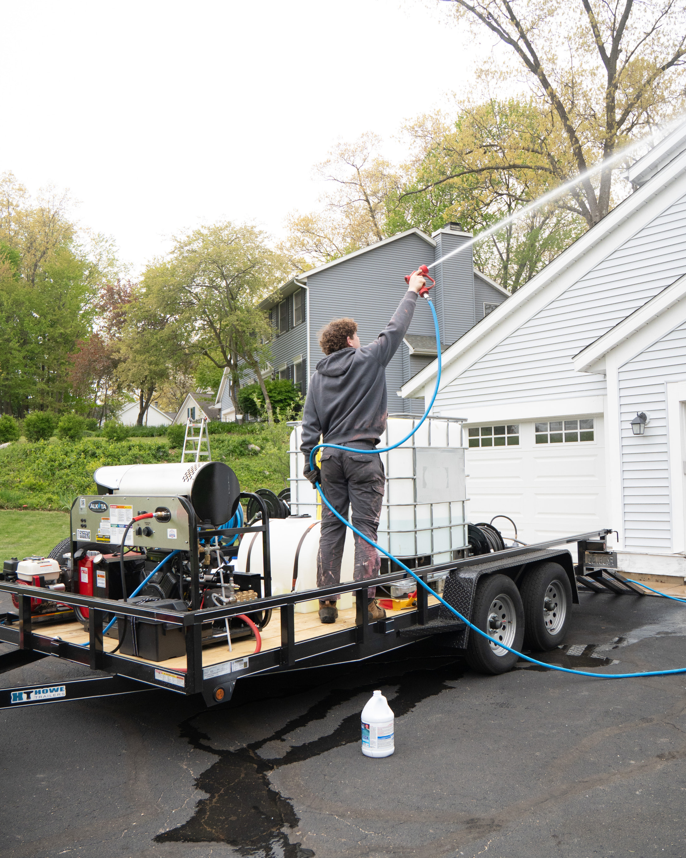 Technician soft-washing a roofline from the trailer rig, with pressure washer, water tank and hose reel aboard